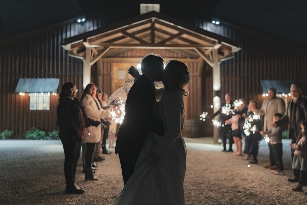 Rae & David kiss during a sparkler send-off outside the lit barn entrance at Covey Creek at night