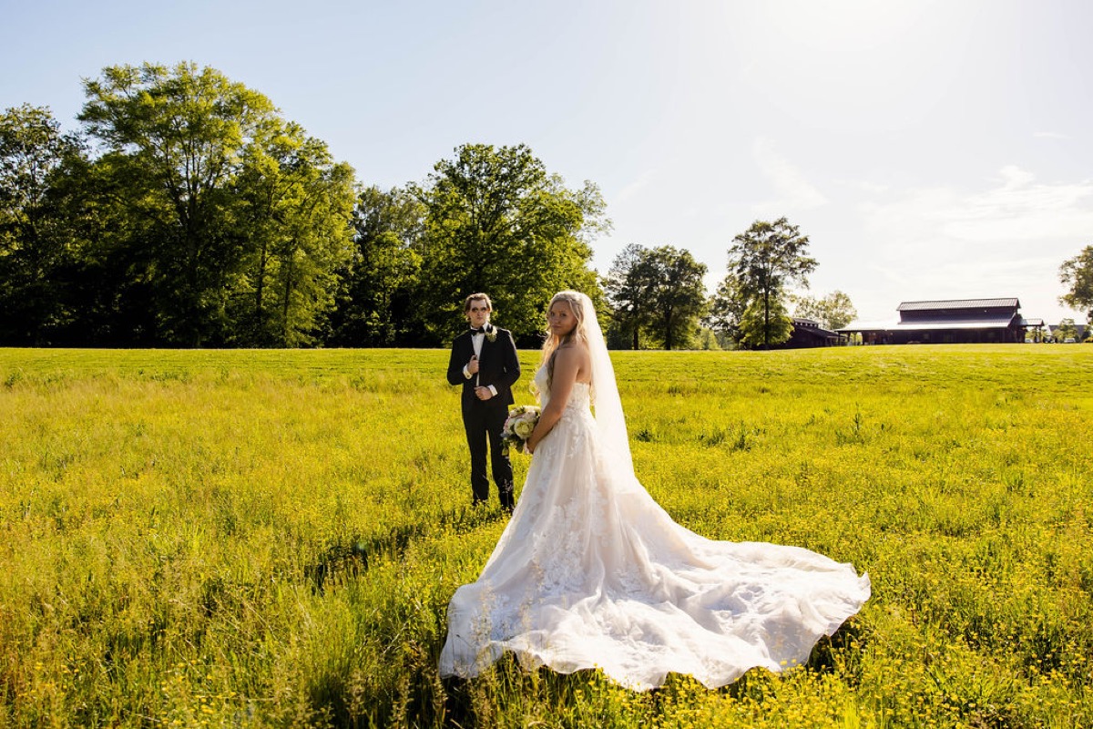 Raven & Jackson pose in tall grass, her lace train spread, with Covey Creek's barn behind them