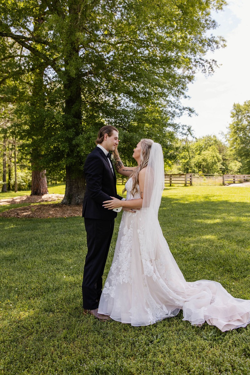 Raven & Jackson stand close under a leafy tree on the green lawn at Covey Creek, her hand on his cheek