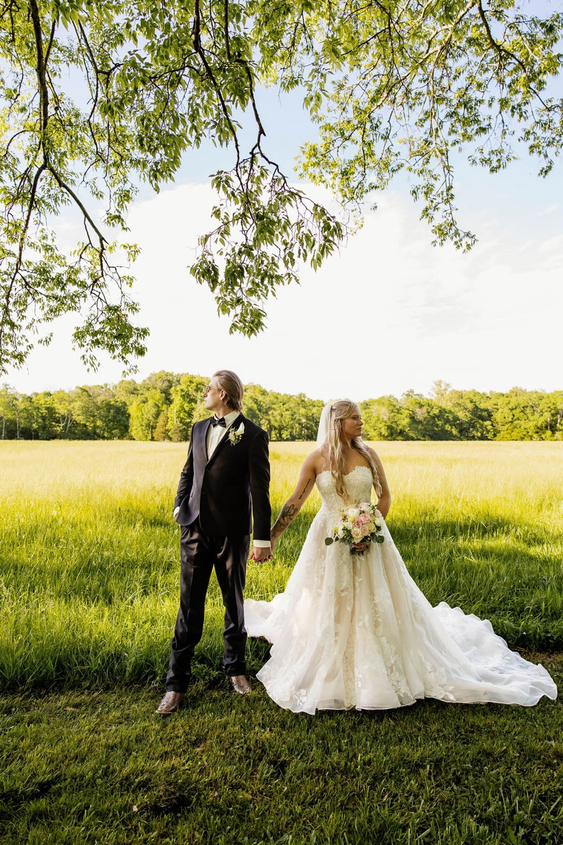Raven & Jackson hold hands looking opposite ways in a sunlit grassy field under trees at Covey Creek
