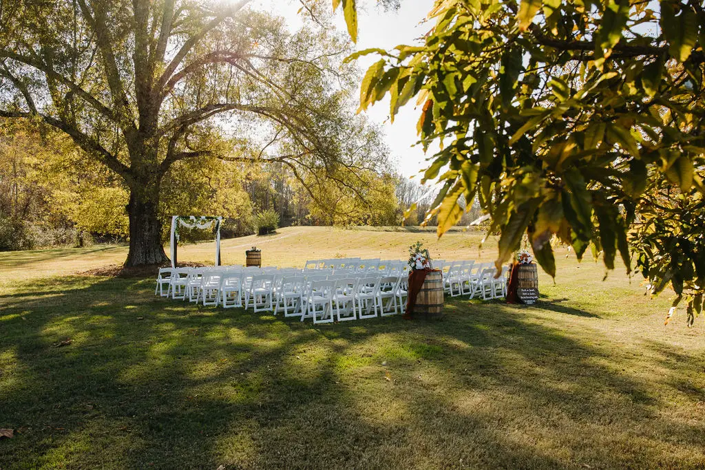Outdoor ceremony arbor under autumn trees with white chairs and whiskey-barrel florals for Rebecca & Troy at Covey Creek.