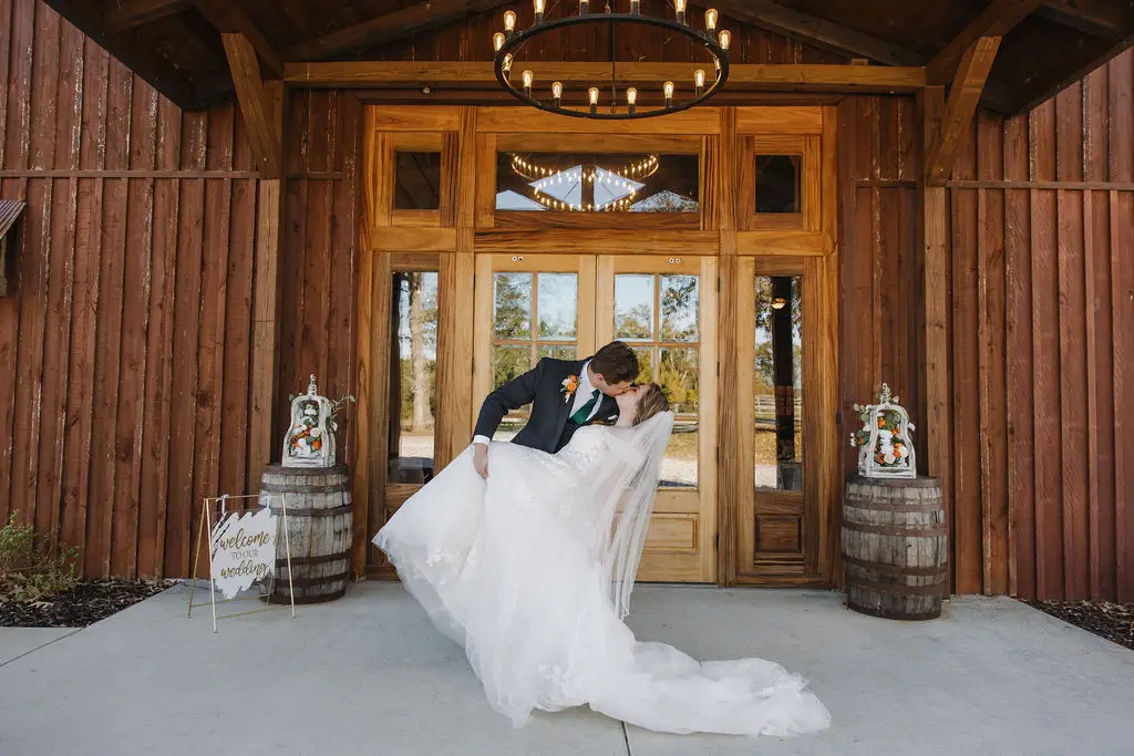Rebecca & Troy share a dip kiss at Covey Creek’s rustic barn doors, flanked by whiskey barrels and florals.