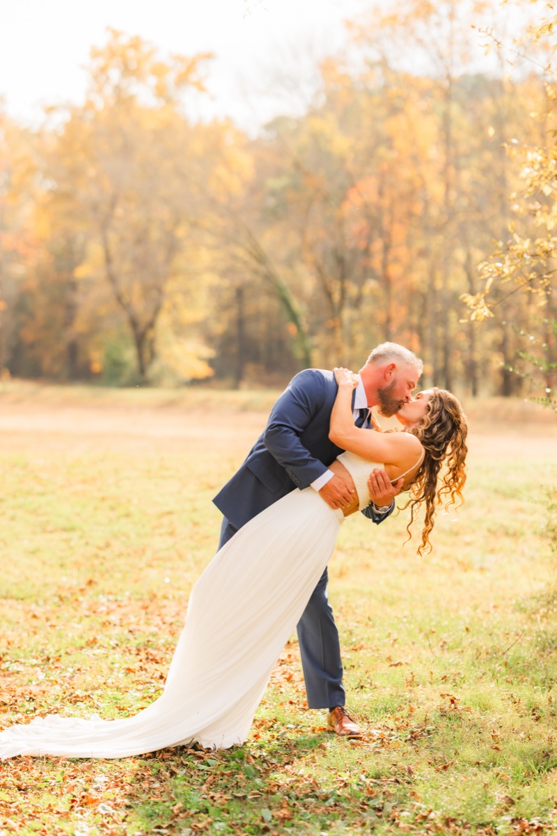 Sarah & Adam share a dip kiss in a sunny field before golden fall trees at their Covey Creek wedding