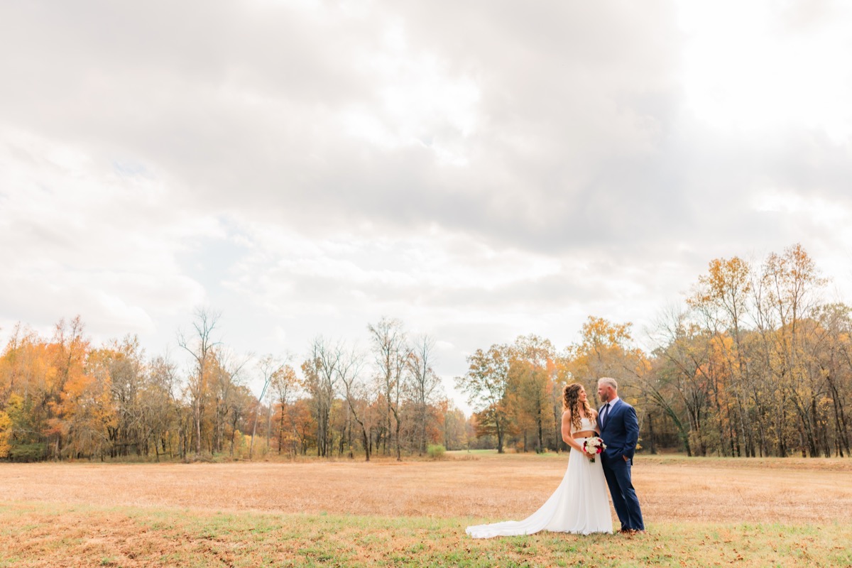 Sarah & Adam stand together in a golden autumn field with her red-and-white bouquet at Covey Creek