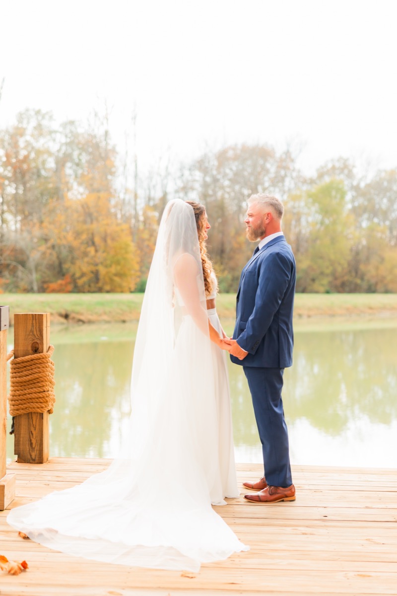 Sarah & Adam hold hands on the wooden Pond dock with autumn trees across the water at Covey Creek