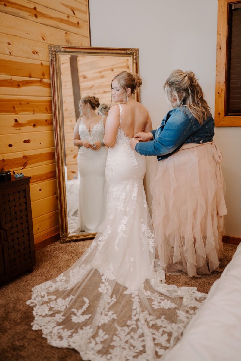 Shay & Wyatt's day begins as Shay's open-back lace gown is fastened before a gold mirror at Covey Creek.