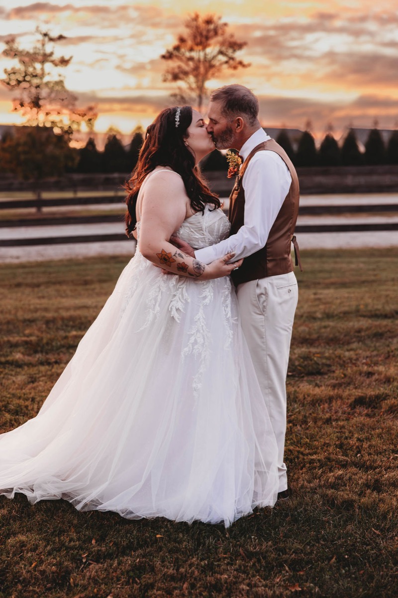 Tori & Tim share a kiss in an open grassy field at sunset with a wooden fence at Covey Creek