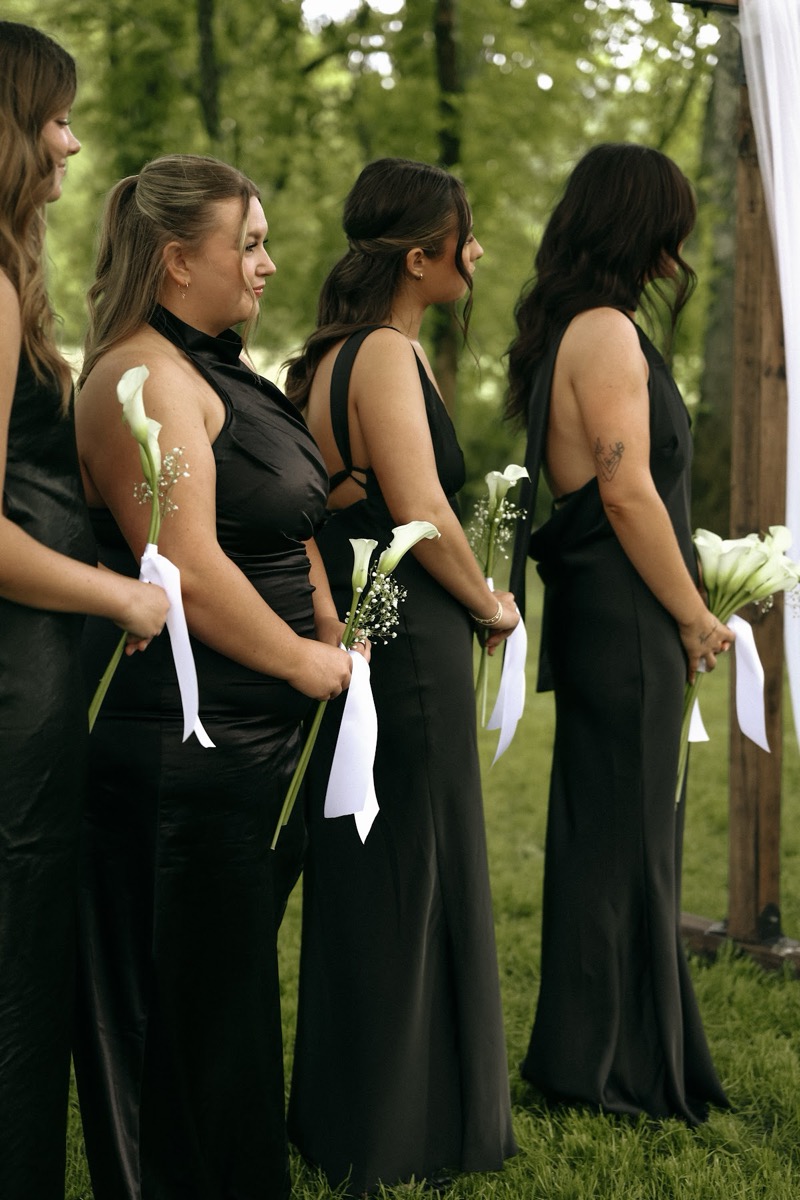 Bridesmaids in black hold white calla lilies beside Cadence & Brody's wood-arch ceremony at Covey Creek