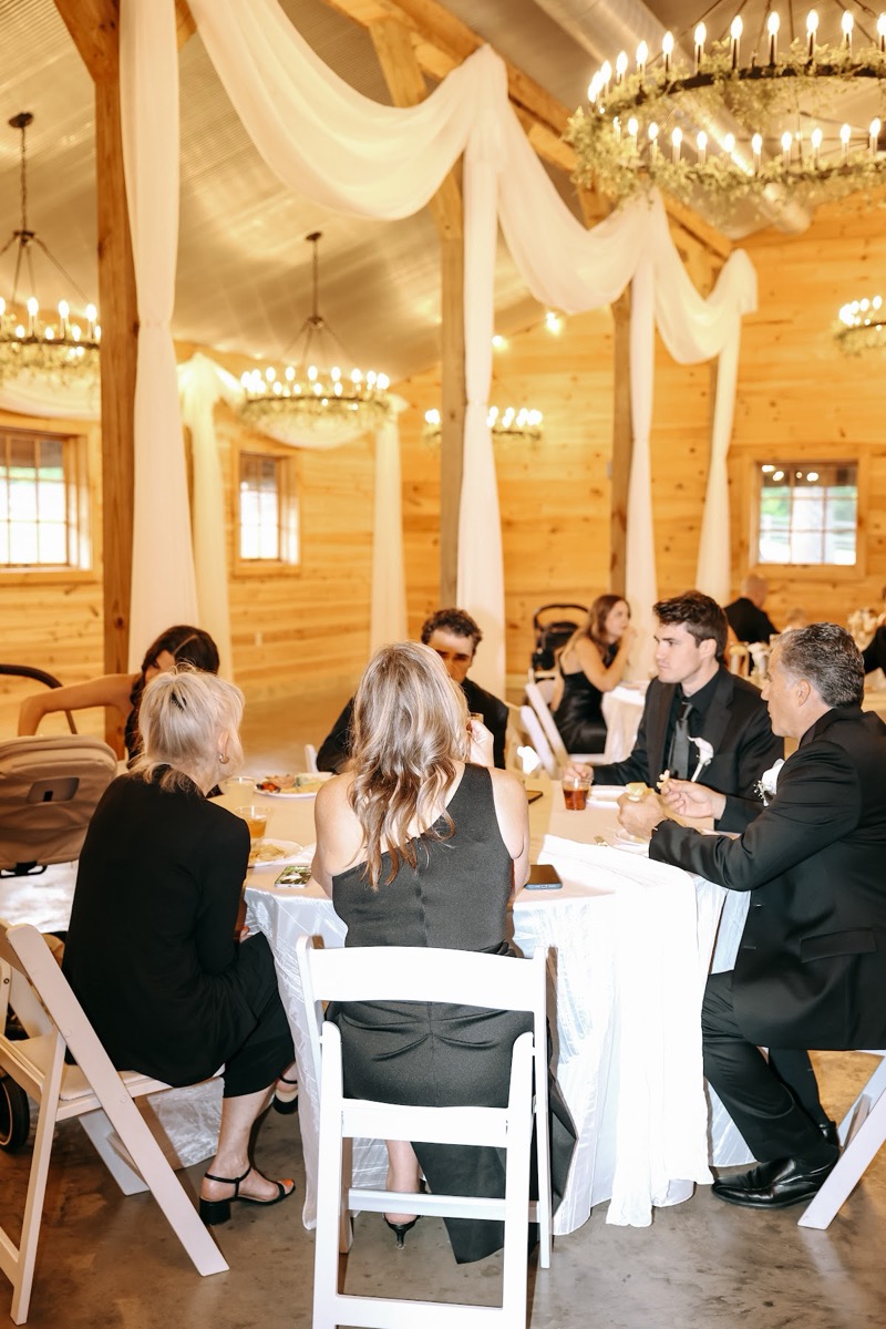 Guests in black dine under iron chandeliers in the barn at Cadence & Brody's Covey Creek reception
