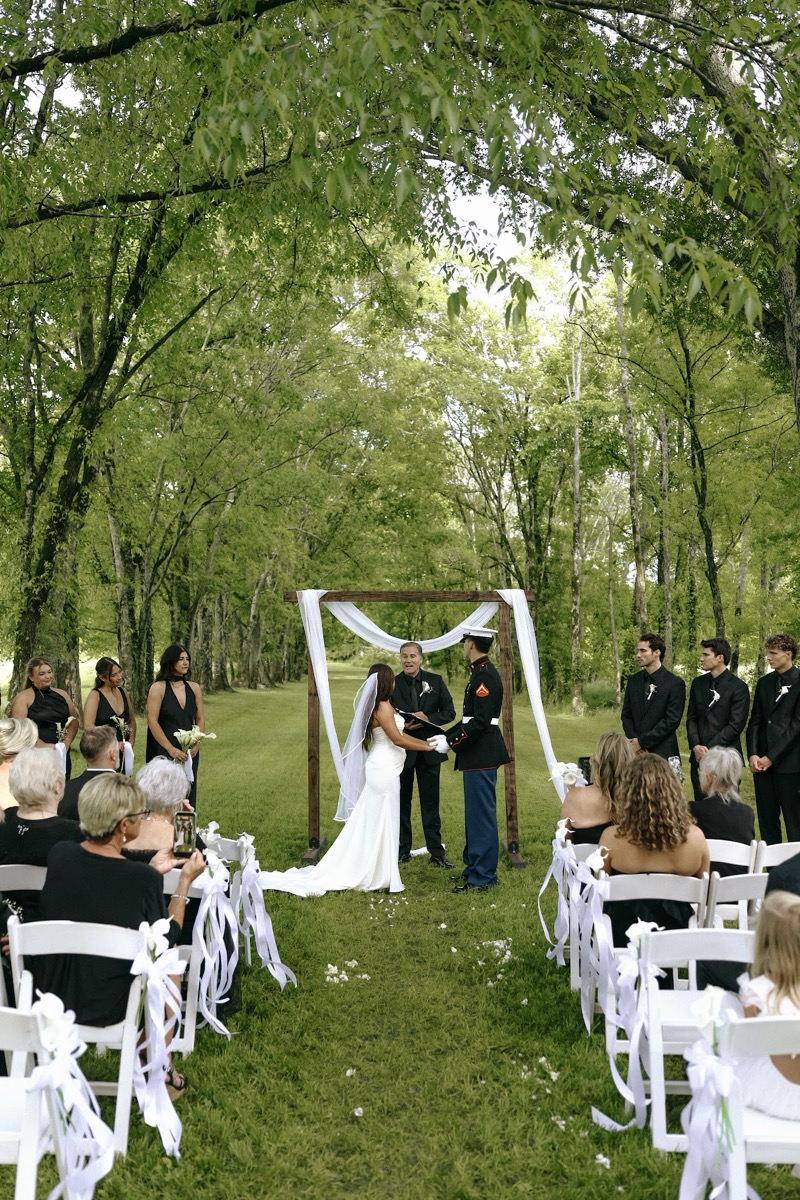Cadence & Brody stand under a fabric-draped wood arch on the tree-lined lawn as guests watch at Covey Creek