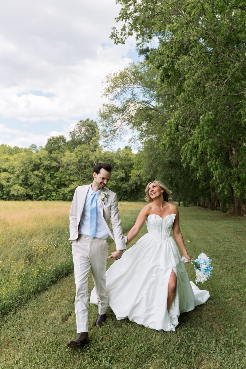 Kyley & Nick walk hand in hand past meadow grass and trees after their ceremony at Covey Creek