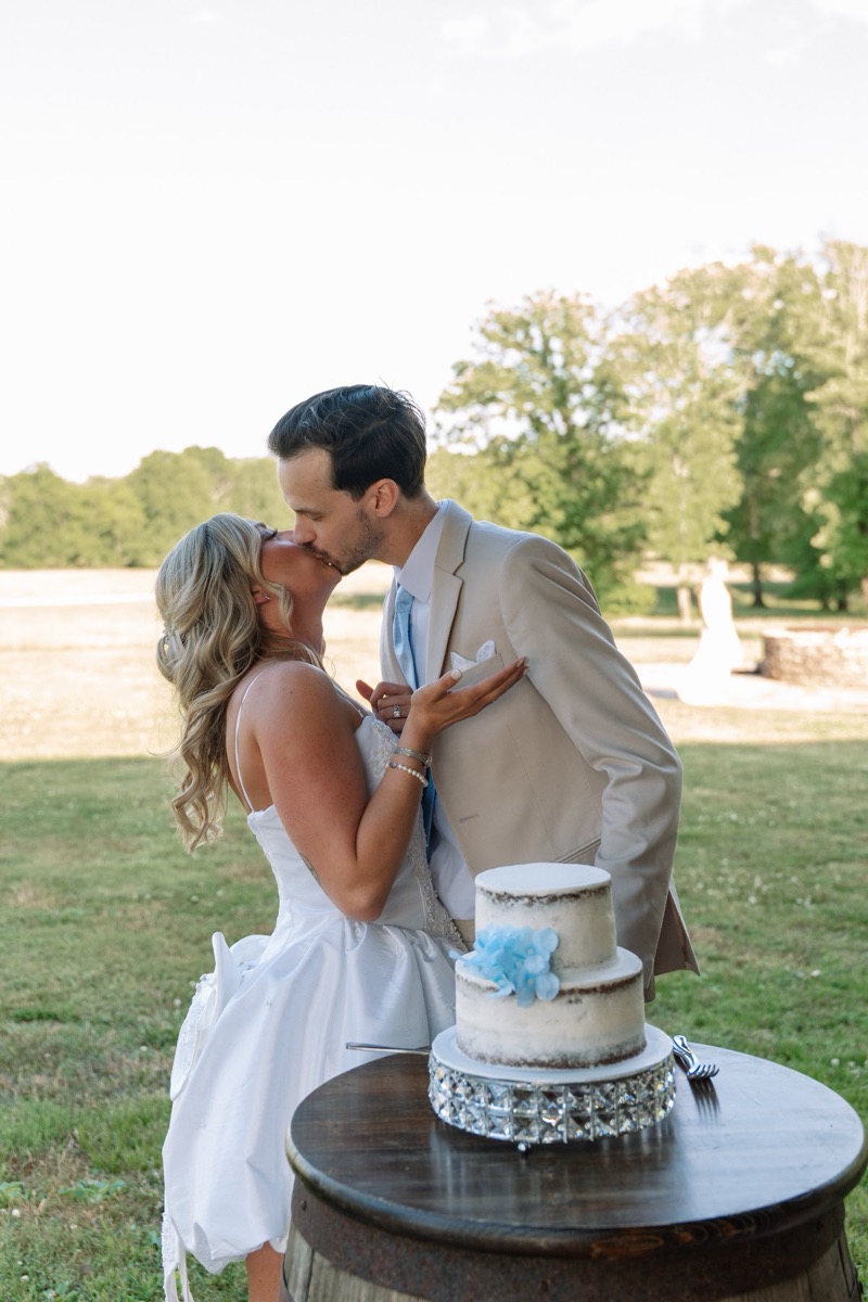 Kyley & Nick kiss behind their blue-flower cake on a barrel on the outdoor lawn at Covey Creek