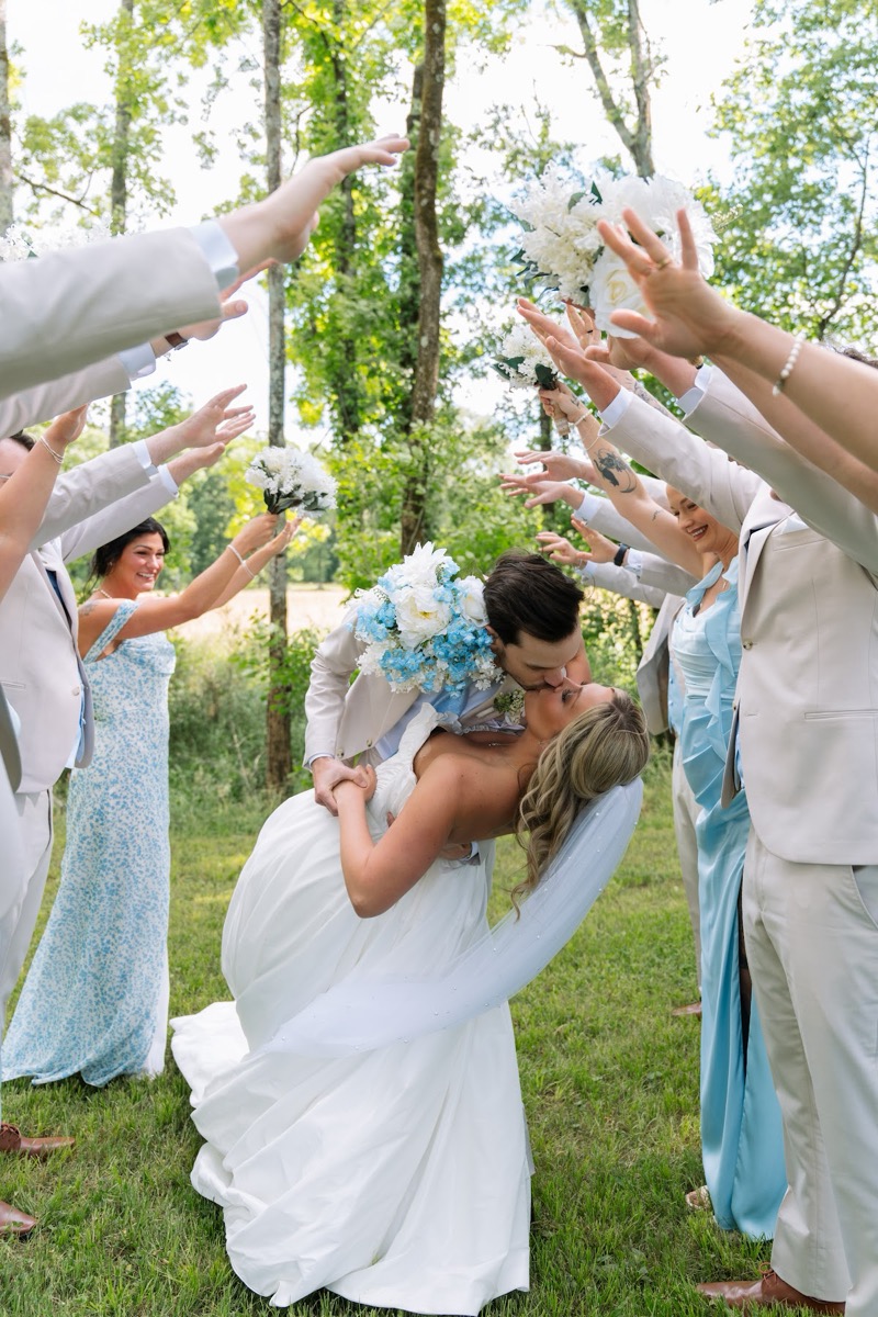 Kyley & Nick kiss under an arch of raised arms from their wedding party on the lawn at Covey Creek