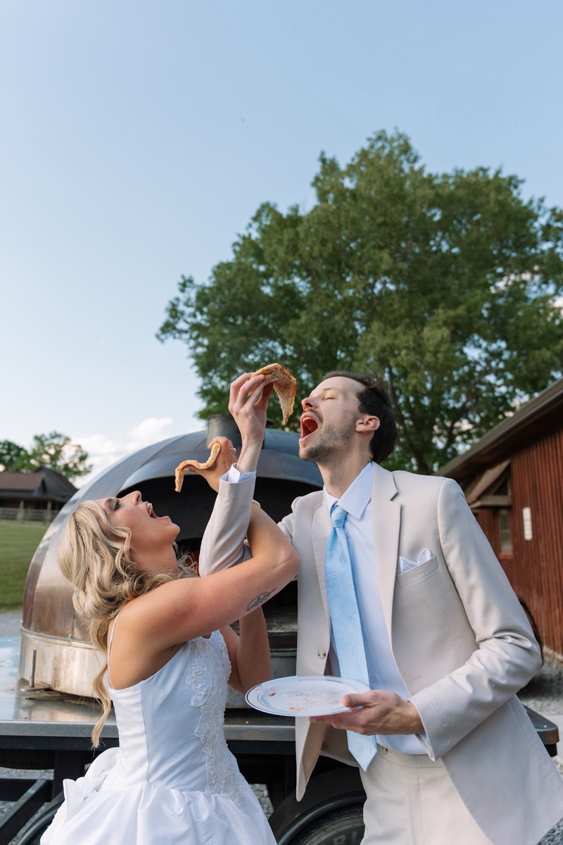 Kyley & Nick playfully feed each other pizza beside a wood-fired oven outdoors at Covey Creek