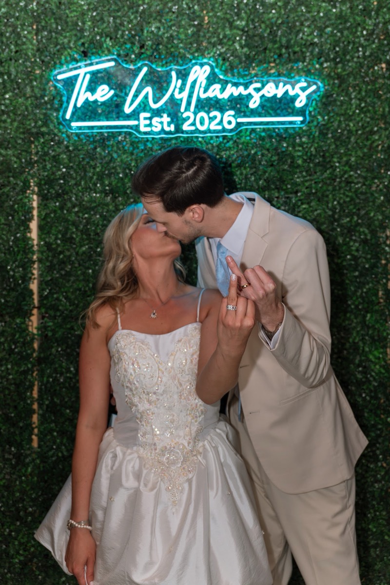 Kyley & Nick kiss playfully under a neon Williamsons Est. 2026 hedge-wall sign at Covey Creek