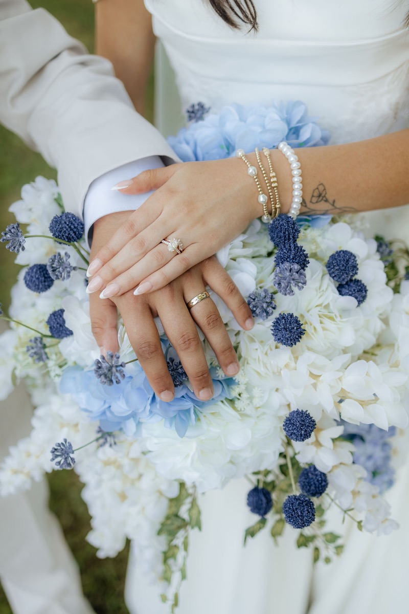 Kylie & Mason show wedding rings on a white and blue bouquet against spring grass at Covey Creek