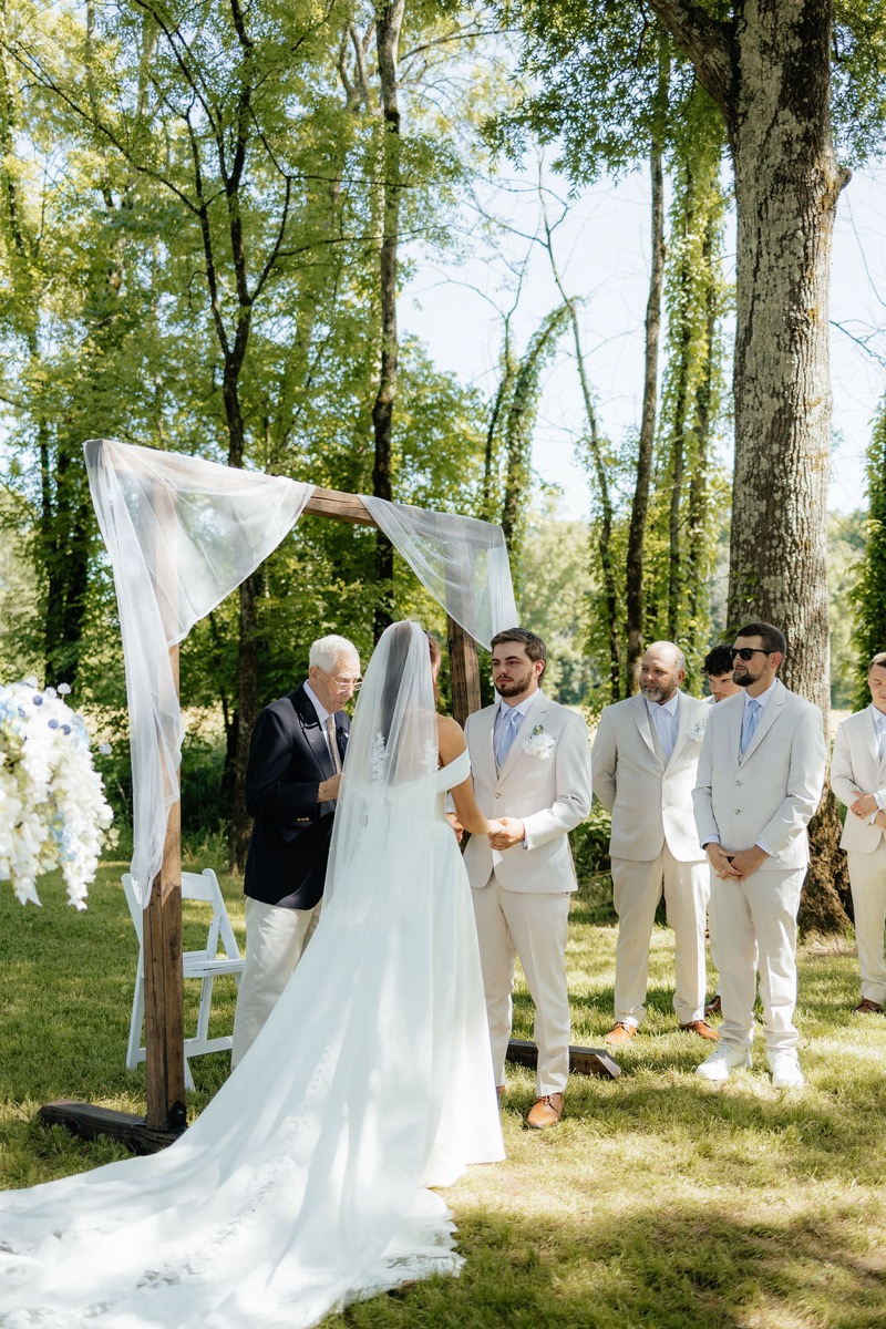 Kylie & Mason hold hands under a white-draped arbor during their Tunnel of Love ceremony at Covey Creek