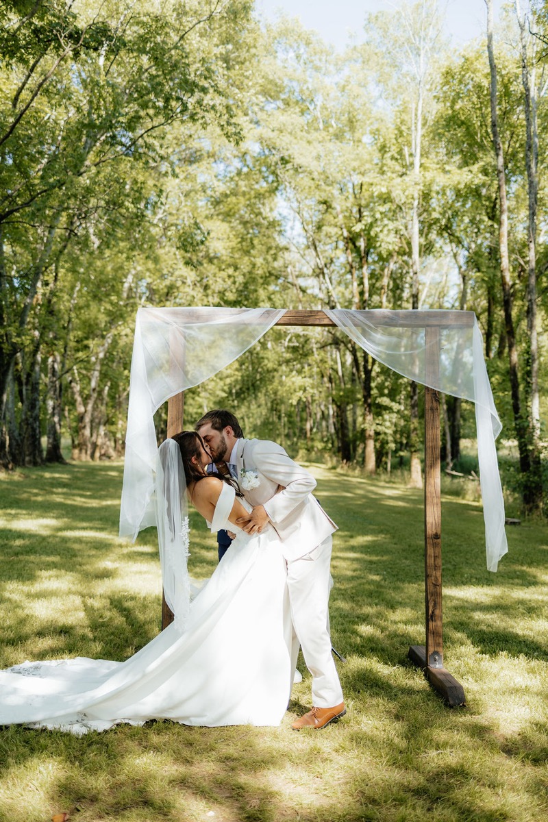 Kylie & Mason share a dip kiss under a wooden arbor on a sunny Tunnel of Love lawn at Covey Creek