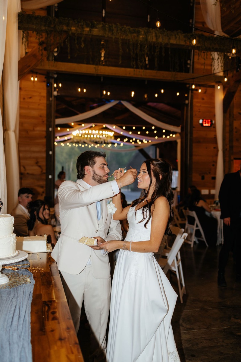 Kylie & Mason feed each other cake under string lights at their Covey Creek indoor reception