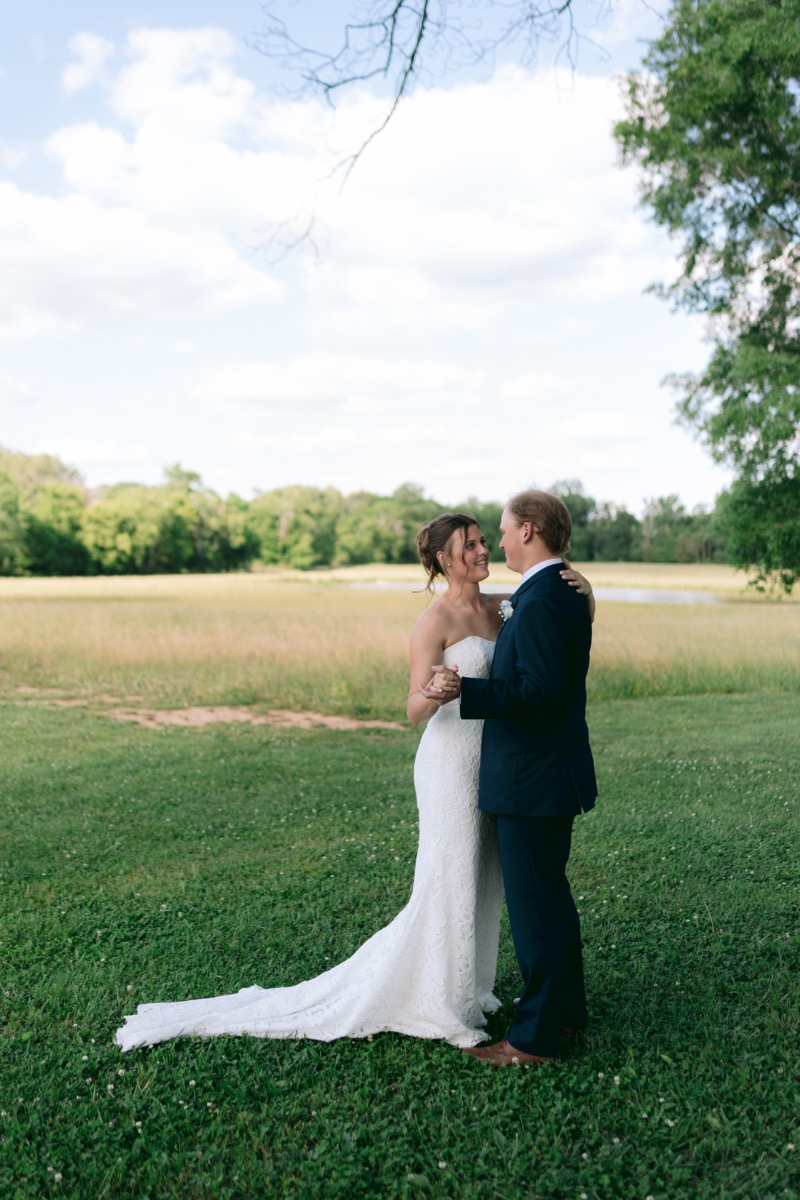 Rachel & Ben smile hand-in-hand by the Pond with lush spring trees behind them at Covey Creek.
